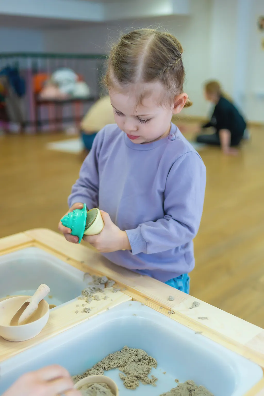 A toddler in a lilac jumper fills a sand mould at a wooden sand and water table