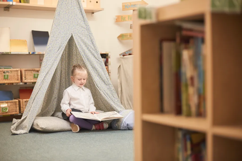 A young girl sits cross legged on cushions in a fabric reading tent, turning the pages of a picture book