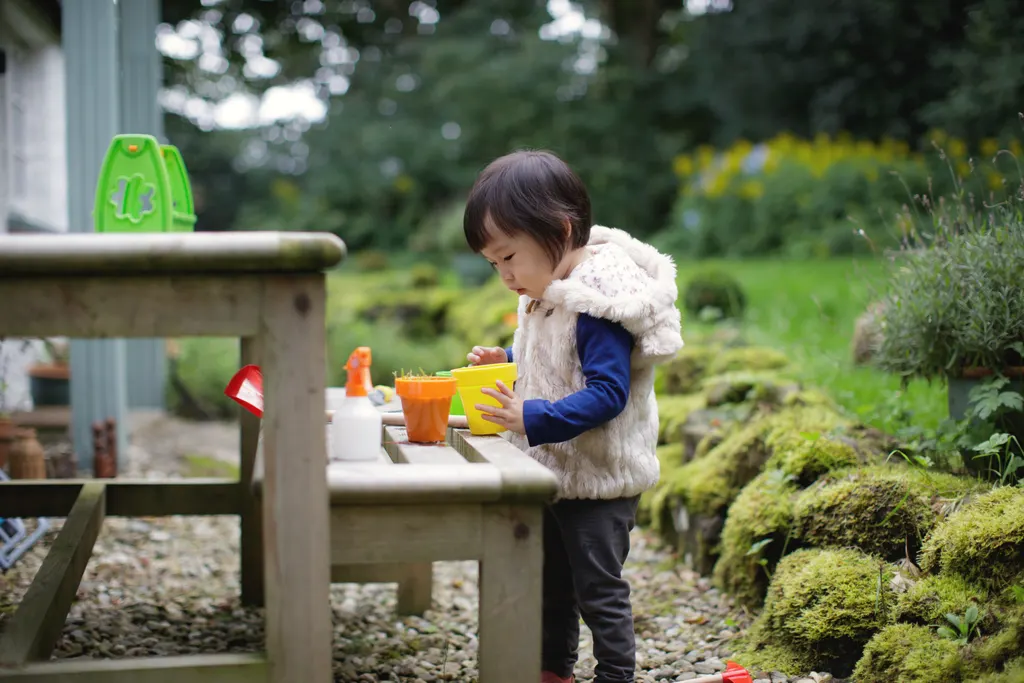 A toddler in a fleece gilet plants seedlings in small pots at a wooden outdoor play kitchen beside a mossy garden bed